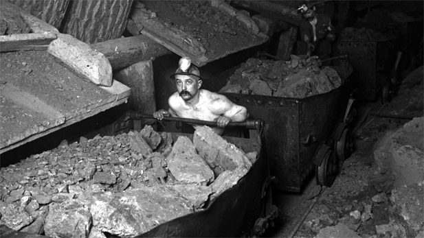 A mile underground, a shirtless trammer pushes a car filled with rock as two men filling cars in the background. Calumet-Hecla Mines, Calumet, Michigan.