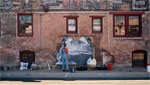 Two women place art on dirtied brick wall.