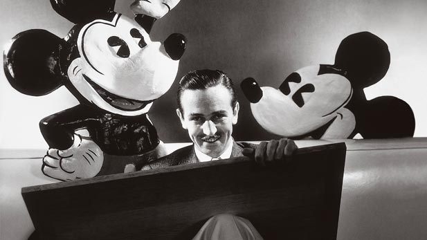Walt Disney seated with drawing board on his lap and representations of his creations Mickey and Minnie Mouse behind. October 1933.