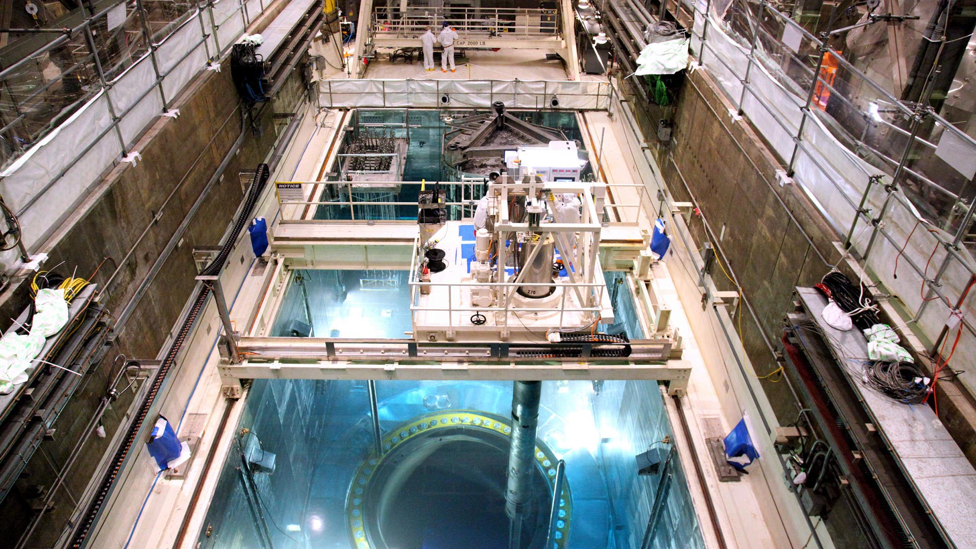 The deep blue of Cherenkov radiation reveals the location of the fuel assemblies within the open reactor core (foreground). The tube beneath the platform, a tube-shaped fuel handling machine extracts one of these assemblies.