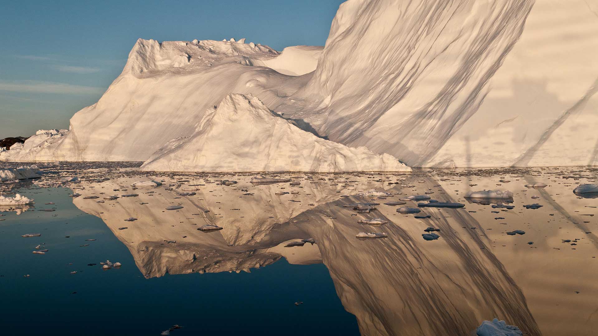 The midnight sun casts a golden glow on an iceberg and its reflection in Disko Bay, Greenland. Much of Greenland’s annual mass loss occurs through calving of icebergs such as this.