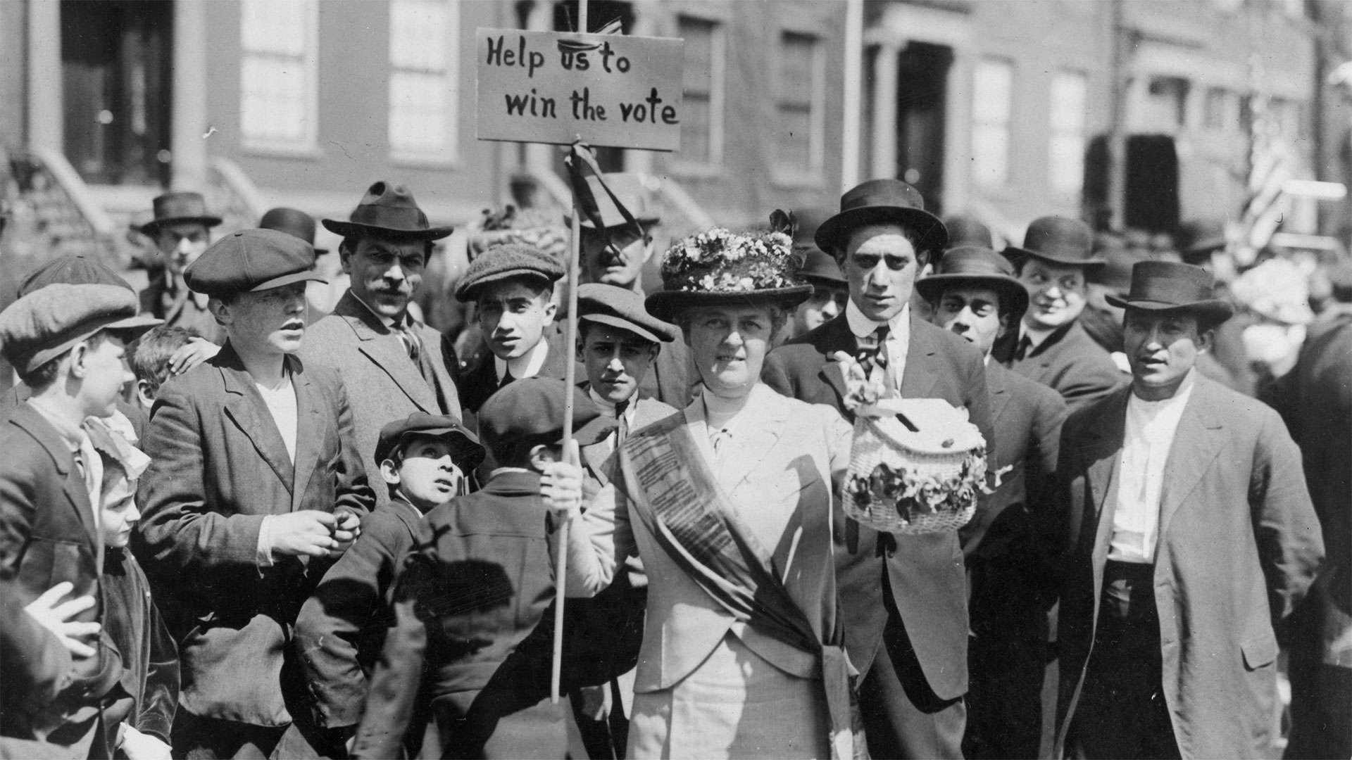 Suffragist, "Mrs. Suffern," holding sign; crowd of boys and men behind. New York, 1914.