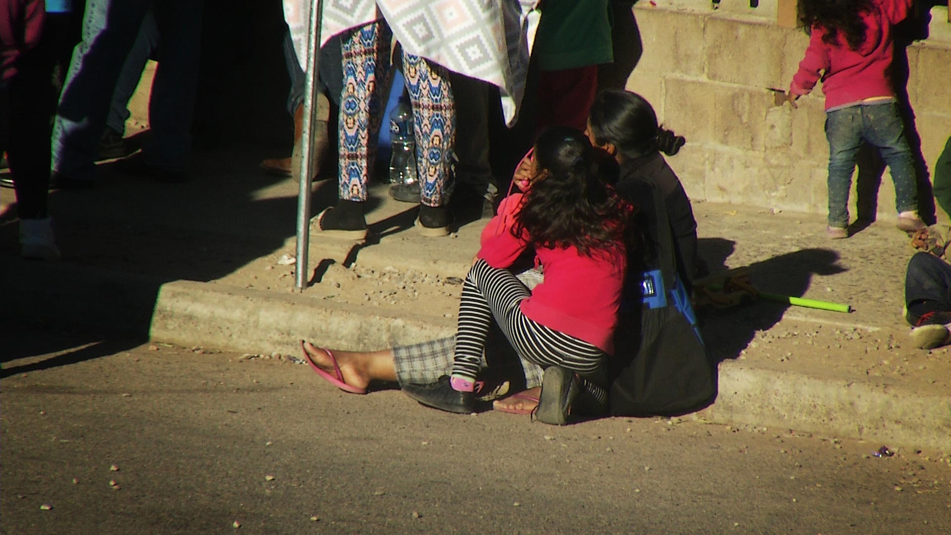 A woman and girl sit on the sidewalk with a group of migrants as they wait to enter the Kino Border Initiative's cafeteria. The nonprofit provides humanitarian aid to migrants and deportees in Nogales, Sonora. February 2020.