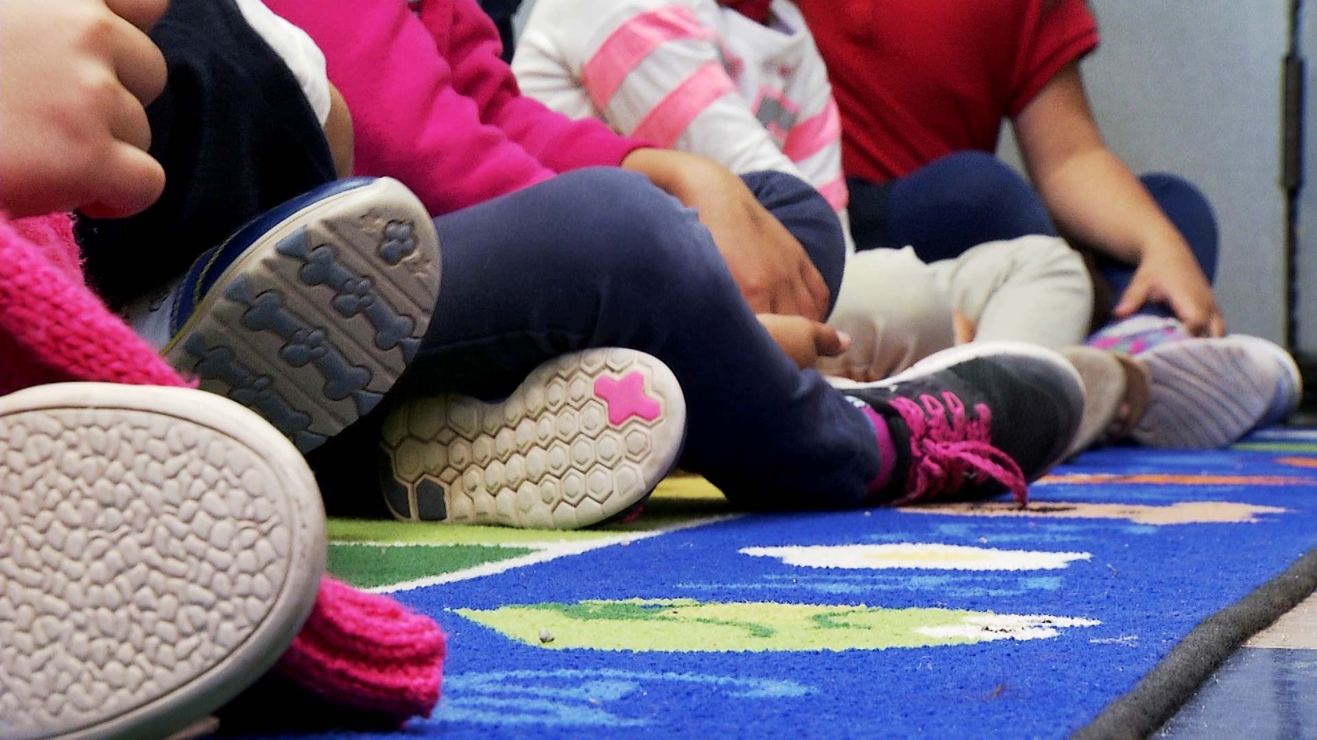 The shoes of children as they sit in a row inside a kindergarten classroom. 