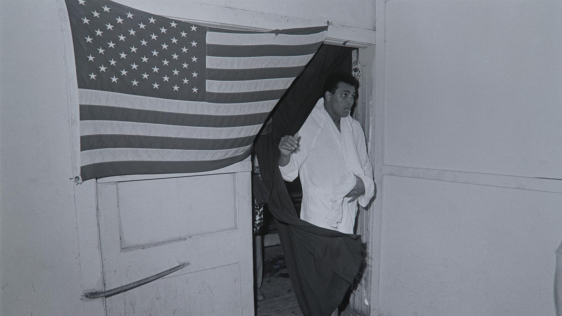 Muhammad Ali coming through doorway draped in a United States flag. Fifth Street Gym. Miami, FL. February 25, 1971.