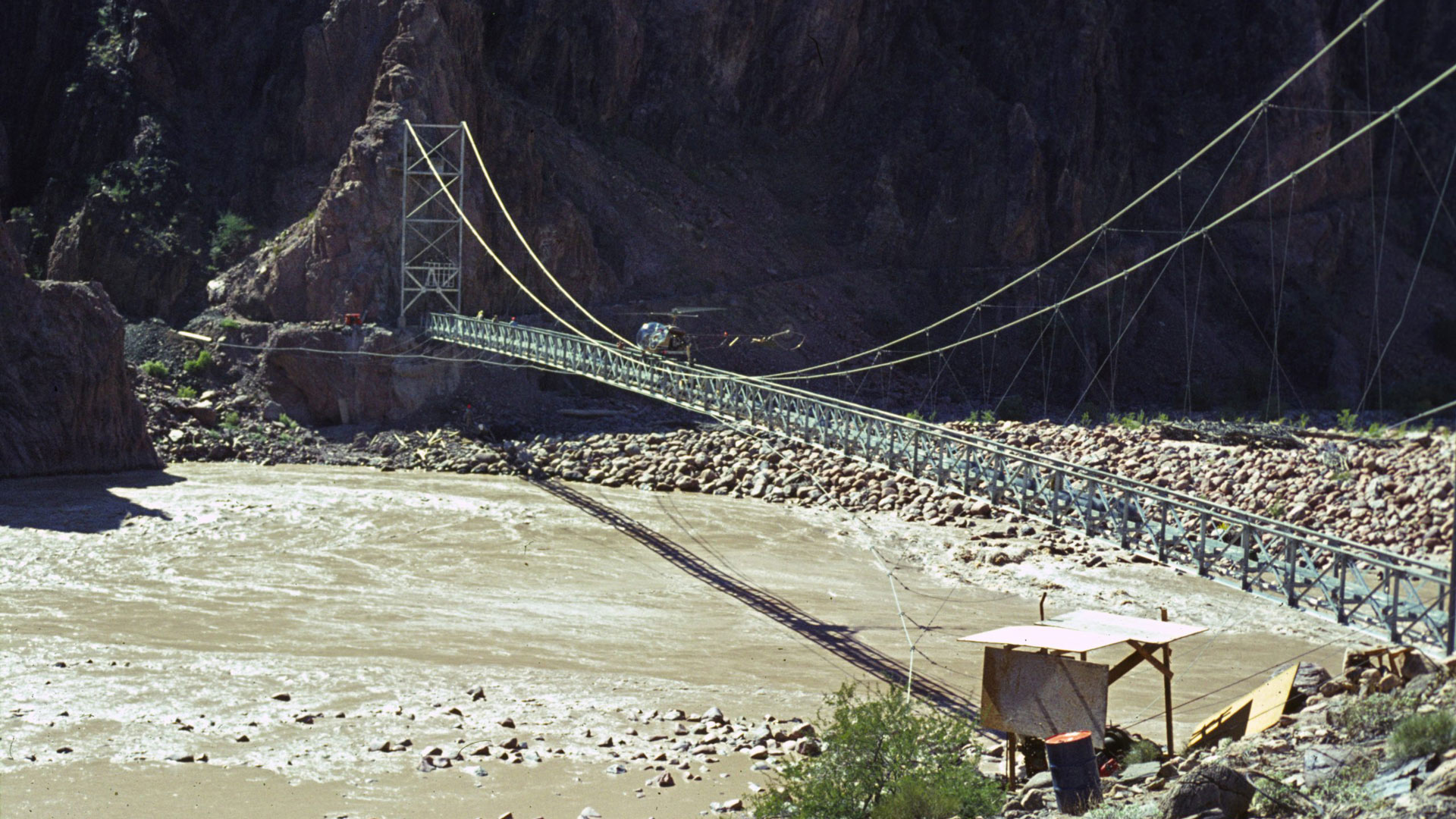 Construction on the Trans-Canyon Waterline started in 1965. This photo shows a contractor’s helicopter delivering materials to attach the pipe beneath the Silver Bridge over the Colorado River.