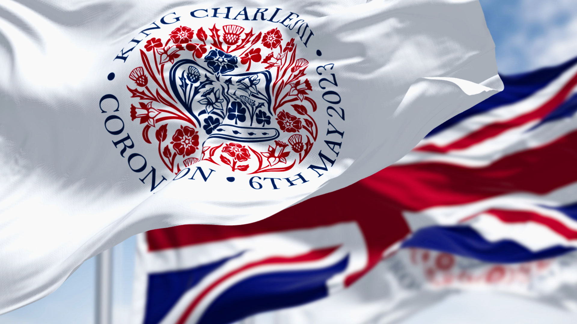 Flags with the emblem of the coronation of King Charles III and of UK waving. The emblem depicts flora of the four nations in the shape of St Edward’s Crown.